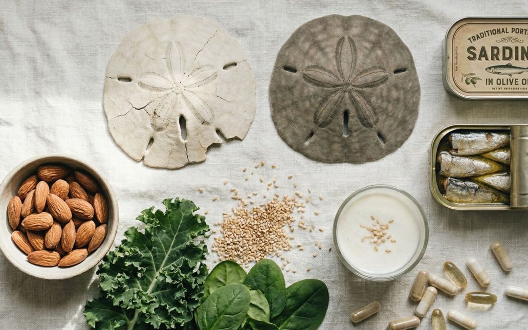A flat lay of two sand dollars — one bleached and cracked, one intact — alongside bone-supporting foods including almonds, kale, spinach, sardines, sesame seeds, kefir, and supplements on a linen background