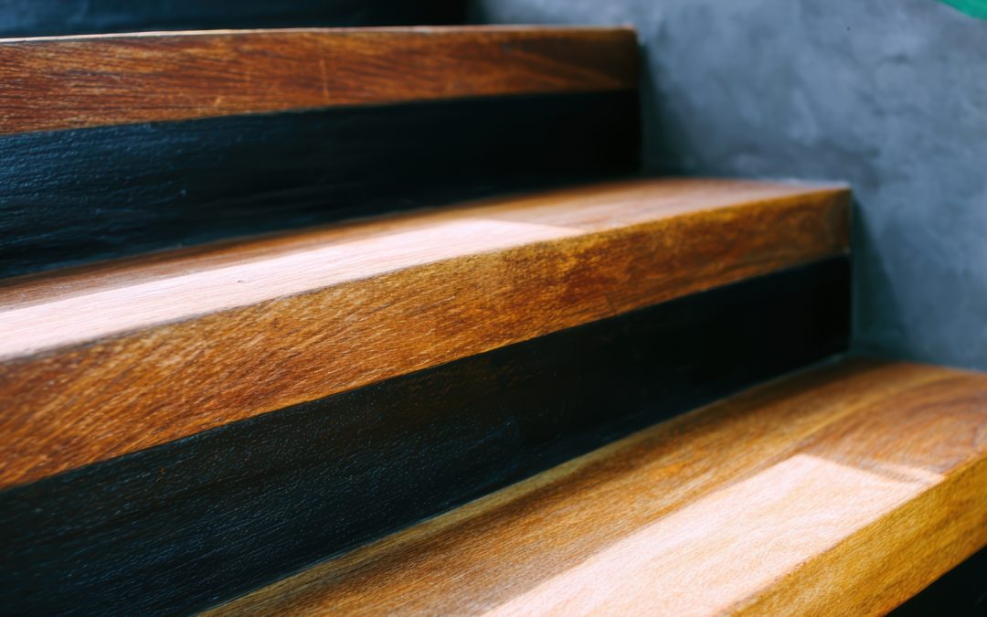 A close-up of a worn wooden staircase with visible grain and scuff marks, sunlight casting shadows across the steps, a teal railing visible in the background.