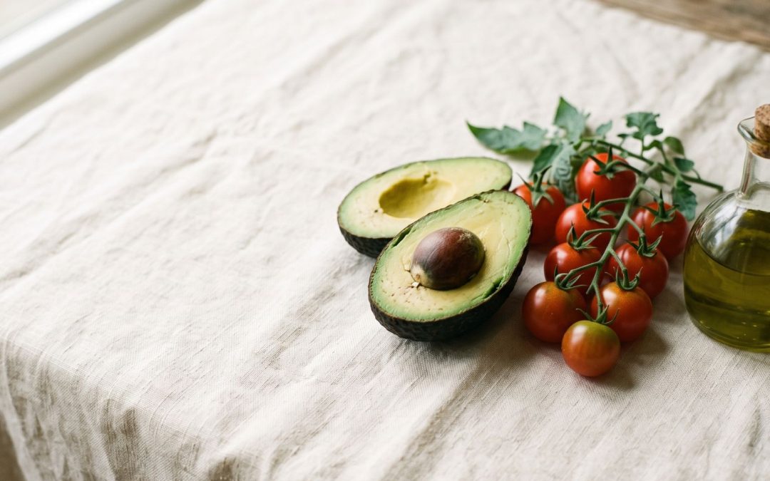 Halved avocado, vine cherry tomatoes and a glass cruet of olive oil on cream linen — a simple food pairing for better nutrient absorption.