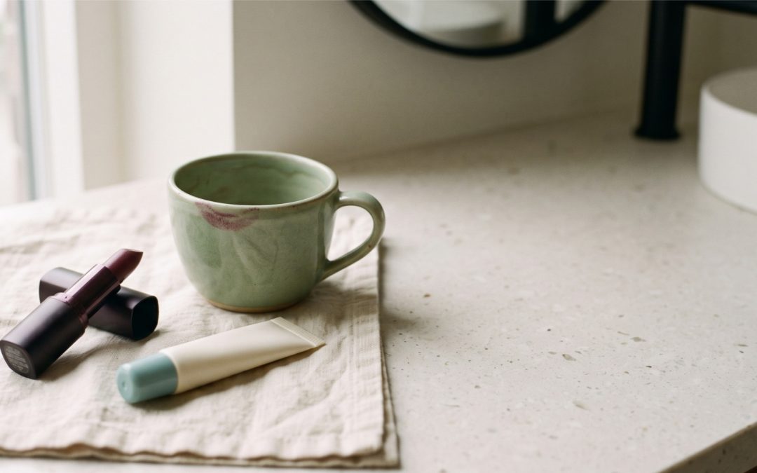A plum satin lipstick and an unbranded cream balm tube on linen beside a coffee cup, for an article on lip health